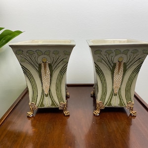 May include: A pair of square ceramic planters with a cream-coloured base, featuring green and gold floral patterns on each side. Each planter is supported by four ornate gold-toned metal feet shaped like claws, providing a raised, stable base.