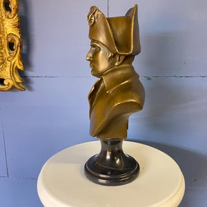 K&ouml;nnte beinhalten: Eine bronzefarbene B&uuml;ste einer historischen Figur, wahrscheinlich Napoleon, mit detailliertem Hut und Mantel. Die B&uuml;ste ist auf einem schwarzen Marmorsockel montiert und steht auf einer wei&szlig;en Oberfl&auml;che. Der Hintergrund ist eine blaue Wand.