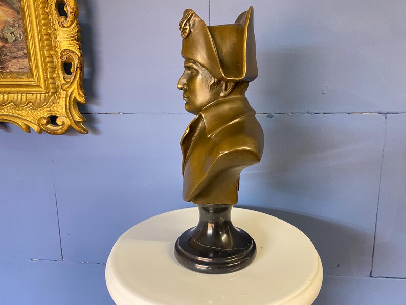 K&ouml;nnte beinhalten: Eine bronzefarbene B&uuml;ste einer historischen Figur, wahrscheinlich Napoleon, mit detailliertem Hut und Mantel. Die B&uuml;ste ist auf einem schwarzen Marmorsockel montiert und steht auf einer wei&szlig;en Oberfl&auml;che. Der Hintergrund ist eine blaue Wand.