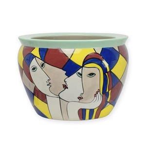 Puede incluir: Una maceta de cerámica con un borde verde claro y un diseño geométrico colorido. La maceta presenta una ilustración estilizada de tres caras con labios rojos, cabello azul y formas geométricas amarillas y azules.