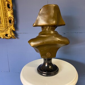 K&ouml;nnte beinhalten: Bronzeb&uuml;ste einer historischen Figur, m&ouml;glicherweise Napoleon, mit einem Zweispitzhut. Die Skulptur ist auf einem schwarzen Marmorsockel montiert und steht auf einer wei&szlig;en Oberfl&auml;che. Die B&uuml;ste hat ein detailliertes Gesicht und Schultern.