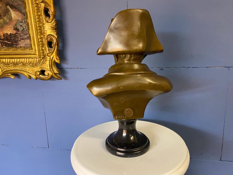 K&ouml;nnte beinhalten: Bronzeb&uuml;ste einer historischen Figur, m&ouml;glicherweise Napoleon, mit einem Zweispitzhut. Die Skulptur ist auf einem schwarzen Marmorsockel montiert und steht auf einer wei&szlig;en Oberfl&auml;che. Die B&uuml;ste hat ein detailliertes Gesicht und Schultern.