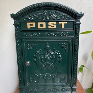 Może przedstawiać: Zielona metalowa skrzynka pocztowa z ozdobnymi detalami i złotym napisem "POST".