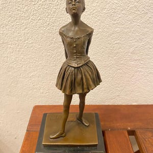 Może przedstawiać: Brązowa rzeźba młodej baletnicy w tutu, stojącej na kwadratowej podstawie. Rzeźba ma szczegółowy gorset i spódnicę, z ramionami baletnicy za plecami. Rzeźba ma około 30 cm wysokości.