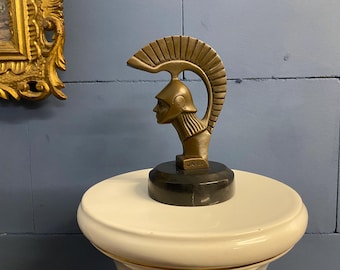 May include: A bronze-colored sculpture of a Roman helmet with a bust of a person in profile. The sculpture is mounted on a black base and sits on a white pedestal. The sculpture has the word "DANET" on the base.