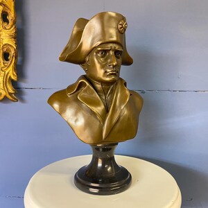 K&ouml;nnte beinhalten: Eine bronzefarbene B&uuml;ste eines Mannes mit Zweispitz und Mantel. Die B&uuml;ste ist auf einem schwarzen Marmorsockel montiert. Die Skulptur wird auf einem wei&szlig;en Tisch vor einer blauen Wand ausgestellt. Im Hintergrund ist ein gerahmtes Gem&auml;lde zu sehen.