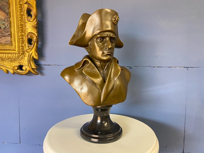 K&ouml;nnte beinhalten: Eine bronzefarbene B&uuml;ste eines Mannes mit Zweispitz und Mantel. Die B&uuml;ste ist auf einem schwarzen Marmorsockel montiert. Die Skulptur wird auf einem wei&szlig;en Tisch vor einer blauen Wand ausgestellt. Im Hintergrund ist ein gerahmtes Gem&auml;lde zu sehen.