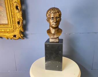 May include: A bronze-colored bust of a man with short hair, mounted on a black marble pedestal. The sculpture is set against a blue wall, with a gold-framed painting visible in the background. The bust is a classical art piece.
