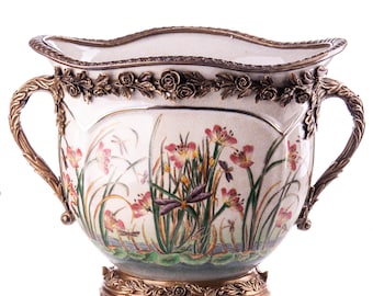 Könnte beinhalten: Ein weißer Keramik-Pflanztopf mit goldenen Akzenten und einem Blumendesign mit rosa Blüten, grünen Blättern und Libellen. Der Topf hat zwei Griffe und einen dekorativen Sockel.