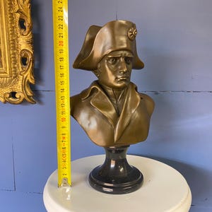 K&ouml;nnte beinhalten: Eine bronzefarbene B&uuml;ste einer historischen Figur, die einen Zweispitz und einen Milit&auml;rmantel tr&auml;gt. Die B&uuml;ste ist auf einem schwarzen Sockel montiert und ist etwa 25 cm hoch. Die B&uuml;ste steht auf einer wei&szlig;en Oberfl&auml;che, mit einem goldgerahmten Gem&auml;lde im Hintergrund.