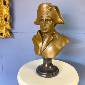 K&ouml;nnte beinhalten: Eine bronzefarbene B&uuml;ste eines Mannes mit Zweispitzhut, auf einem schwarzen Sockel. Die Skulptur zeigt eine historische Figur mit detaillierten Gesichtsz&uuml;gen und Kleidung. Die B&uuml;ste wird auf einer wei&szlig;en Oberfl&auml;che vor einer blauen Wand ausgestellt.