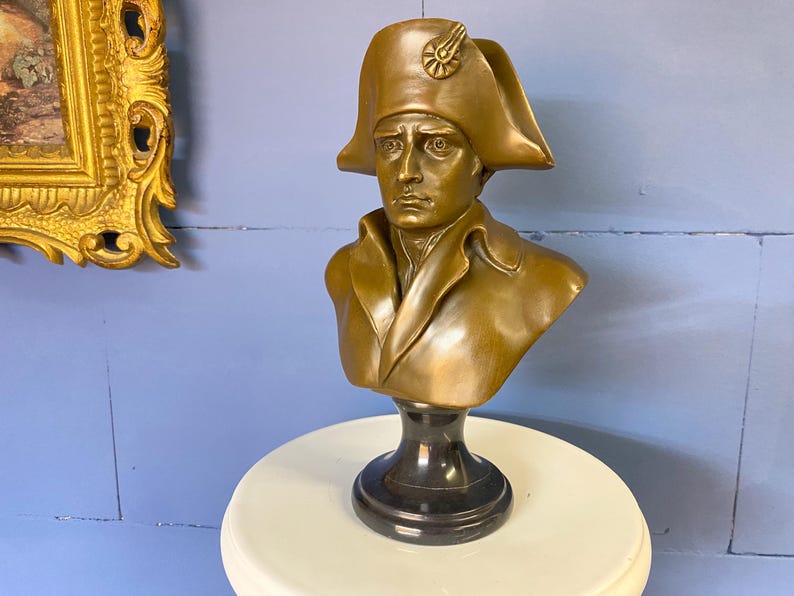 K&ouml;nnte beinhalten: Eine bronzefarbene B&uuml;ste eines Mannes mit Zweispitzhut, auf einem schwarzen Sockel. Die Skulptur zeigt eine historische Figur mit detaillierten Gesichtsz&uuml;gen und Kleidung. Die B&uuml;ste wird auf einer wei&szlig;en Oberfl&auml;che vor einer blauen Wand ausgestellt.