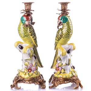 May include: A pair of ornate porcelain parrot candle holders with gold accents. The parrots are perched on branches with flowers and leaves. The candle holders have a gold base with intricate details.