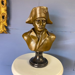 K&ouml;nnte beinhalten: Eine bronzefarbene B&uuml;ste eines Mannes mit Zweispitz und Mantel. Die Skulptur ist auf einem schwarzen Sockel montiert und steht auf einer wei&szlig;en Oberfl&auml;che. Die B&uuml;ste ist eine historische Figur.