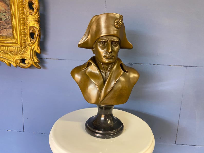 K&ouml;nnte beinhalten: Eine bronzefarbene B&uuml;ste eines Mannes mit Zweispitz und Mantel. Die Skulptur ist auf einem schwarzen Sockel montiert und steht auf einer wei&szlig;en Oberfl&auml;che. Die B&uuml;ste ist eine historische Figur.