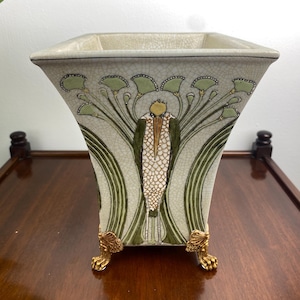 Könnte beinhalten: Eine quadratische, hellfarbene Keramikvase mit ausgestellter Form und dekorativem Blumenmuster in Grün und Gold. Die Vase hat goldfarbene Klauenfüße. Die Vase wird wahrscheinlich zum Präsentieren von Blumen oder Pflanzen verwendet.