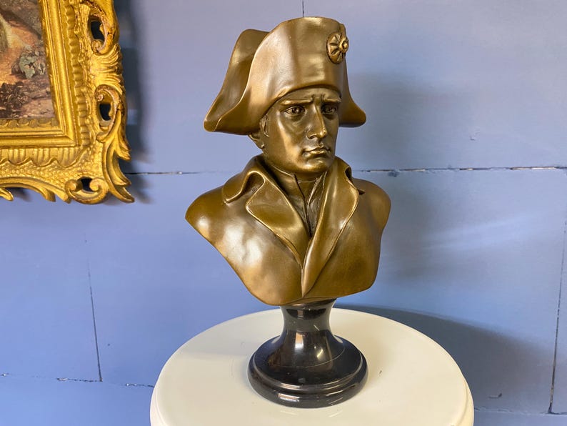 K&ouml;nnte beinhalten: Bronzefarbene B&uuml;ste einer historischen Figur, die einen Mann in einem Mantel und Hut im Milit&auml;rstil zeigt. Die Skulptur ist auf einem schwarzen Sockel montiert und zeigt einen klassischen Stil. Ein dekoratives Objekt.