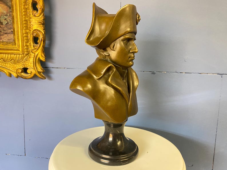 K&ouml;nnte beinhalten: Bronzefarbene B&uuml;ste einer historischen Figur, die einen Mann mit Zweispitz und Milit&auml;rmantel darstellt. Die B&uuml;ste ist auf einem schwarzen Marmorsockel montiert und zeigt ein klassisches Design.