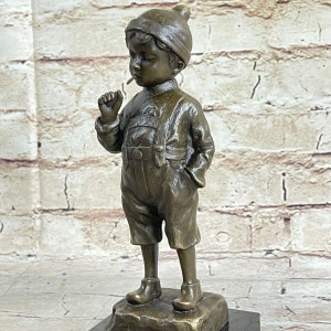 Peut inclure: Une sculpture en bronze d'un jeune garçon portant un chapeau, une salopette et des bottes, avec une sucette à la bouche. Le garçon est debout sur un petit socle texturé. La sculpture a une finition antique détaillée.
