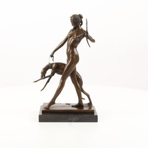 Op de afbeelding: Bronzen sculptuur van een naakte figuur die een windhond aan een lijn uitlaat. De sculptuur is gemonteerd op een rechthoekige basis en heeft een donkerbruine patina. De figuur houdt een lang, dun voorwerp in de andere hand.