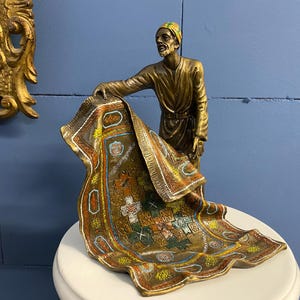 Puede incluir: Una figura de bronce de un hombre sosteniendo una alfombra detallada y colorida. La alfombra presenta intrincados patrones en tonos de rojo, azul y dorado. El hombre lleva un turbante y una túnica larga. La figura está sobre una superficie blanca.