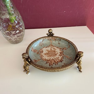 Może przedstawiać: Dekoracyjna miseczka na drobiazgi z niebiesko-brązowym wzorem z porcelany. Miseczka jest podtrzymywana przez trzy nóżki w kształcie cherubina wykonane ze złotego metalu.