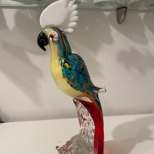 May include: A colourful glass parrot sculpture with a white crest, black beak, and multi-coloured body. The parrot is perched on a clear and red glass branch. The sculpture is displayed on a white surface.