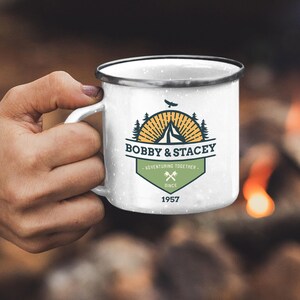 May include: White enamel camping mug with a black rim, held by a hand. The mug features a logo with the text "BOBBY & STACEY," a mountain, trees, and the words "ADVENTURING TOGETHER SINCE 1957." The background shows a campfire.