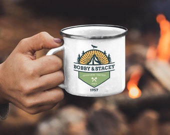May include: White enamel camping mug with a black rim, held by a hand. The mug features a logo with the text "BOBBY & STACEY," a mountain, trees, and the words "ADVENTURING TOGETHER SINCE 1957." The background shows a campfire.