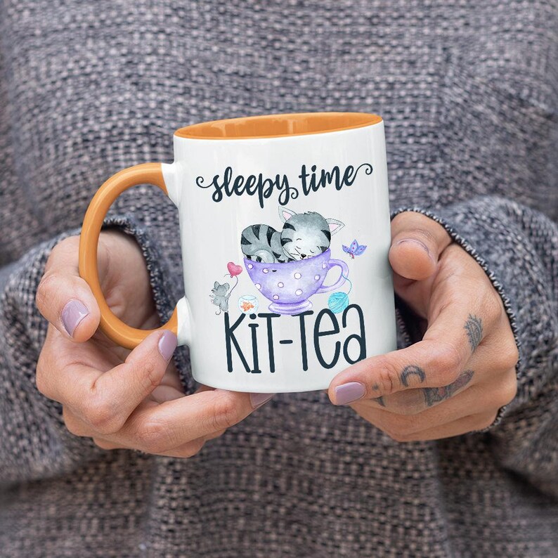May include: White ceramic mug with an orange handle and interior. The mug features a whimsical illustration of a sleeping gray cat in a teacup, with the words "sleepy time" and "Kit-Tea". A cute gift for cat lovers.