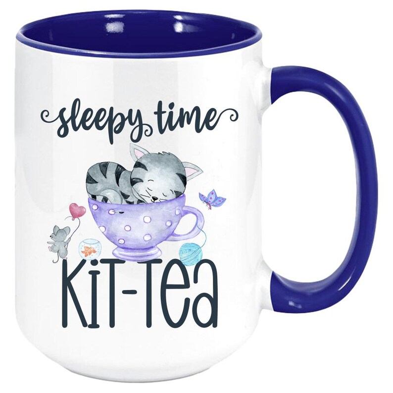 May include: White ceramic mug with a blue interior and handle. The mug features a whimsical design with the words "sleepy time" and "Kit-Tea". A gray tabby cat is sleeping in a purple teacup, with a mouse, butterfly, and yarn balls.