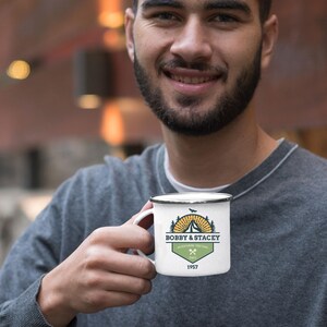 May include: White enamel camping mug with a black rim. The mug features a logo with the text "BOBBY & STACEY" above a tent and sun graphic, with the words "ADVENTURE TOGETHER SINCE 1957". The mug is held by a person wearing a gray sweater.