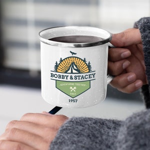 May include: White enamel camping mug with a black rim and speckled finish. The mug features a graphic with the text "BOBBY & STACEY - ADVENTURING TOGETHER SINCE 1957" and a mountain and tree design. The mug is held by a person.