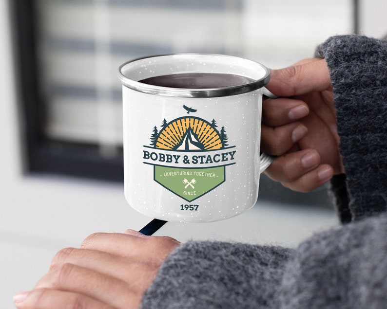 May include: White enamel camping mug with a black rim and speckled finish. The mug features a graphic with the text "BOBBY & STACEY - ADVENTURING TOGETHER SINCE 1957" and a mountain and tree design. The mug is held by a person.