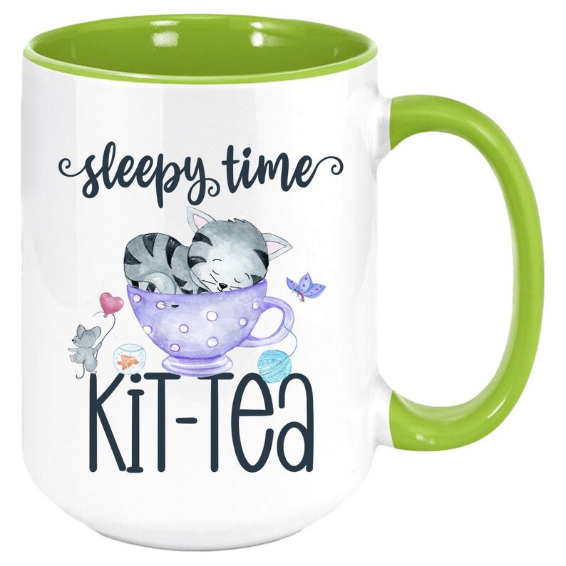 May include: White ceramic mug with a green handle and interior. The mug features a whimsical design with a sleeping gray cat in a purple teacup, a mouse with a heart balloon, and the words "sleepy time kit-tea".