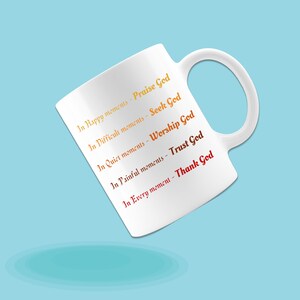 May include: White ceramic mug with a curved handle. The mug features text in orange and red, including "In Happy moments - Praise God" and "In Every moment - Thank God". The mug is set against a light blue background.