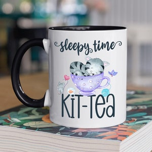 May include: White ceramic mug with a black handle and rim, featuring a whimsical design. The mug has the words "sleepy time" and "Kit-Tea" printed on it. The design includes a sleeping gray cat in a purple teacup, a gray mouse, and a butterfly.