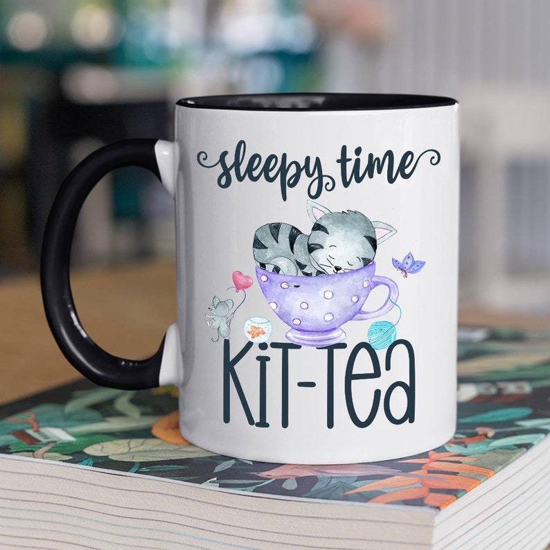 May include: White ceramic mug with a black handle and rim, featuring a whimsical design. The mug has the words "sleepy time" and "Kit-Tea" printed on it. The design includes a sleeping gray cat in a purple teacup, a gray mouse, and a butterfly.