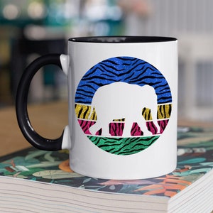 May include: White ceramic mug with a black handle. The mug features a colourful circular design with a white silhouette of an elephant walking on a background of blue, yellow, pink, and green zebra stripes.
