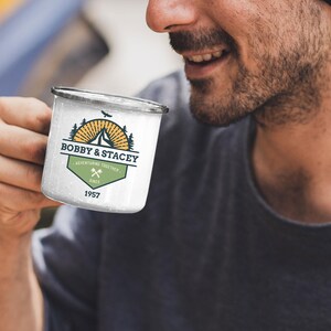 May include: White enamel camping mug with a black rim. The mug features a logo with the text "BOBBY & STACEY ADVENTURING TOGETHER SINCE 1957" with a mountain, sunburst, and crossed axes design. The mug is held by a person.