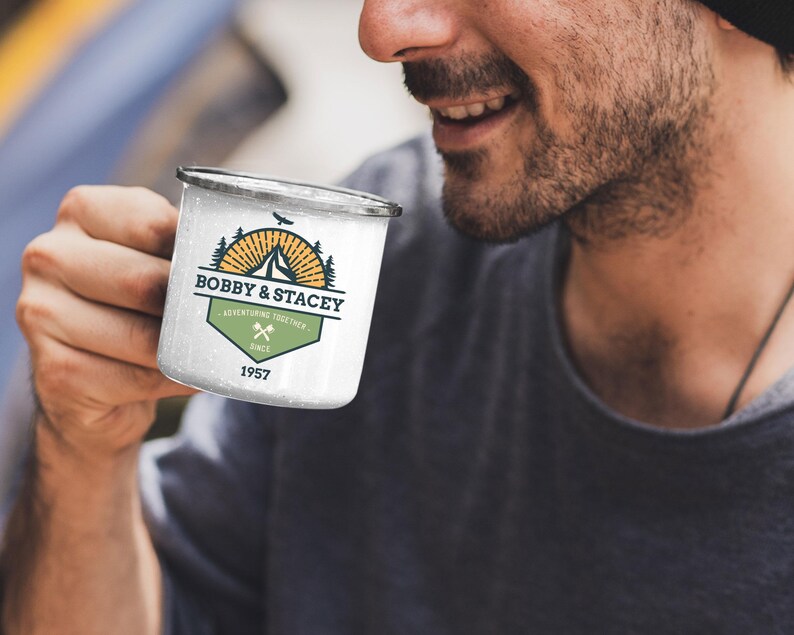 May include: White enamel camping mug with a black rim. The mug features a logo with the text "BOBBY & STACEY ADVENTURING TOGETHER SINCE 1957" with a mountain, sunburst, and crossed axes design. The mug is held by a person.