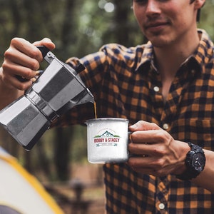 May include: A person pouring coffee from a silver Moka pot into a white enamel mug with a mountain and the text "BOBBY & STACEY SINCE 1998" printed on it.