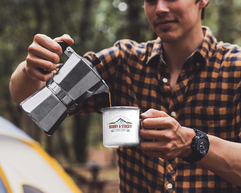 May include: A person pouring coffee from a silver Moka pot into a white enamel mug with a mountain and the text "BOBBY & STACEY SINCE 1998" printed on it.