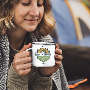 May include: White enamel camping mug with a silver rim. The mug features a green and gold design with the text "BOBBY & STACEY" and "ADVENTURING TOGETHER" with crossed axes and the year "1957". The mug is held by a person.