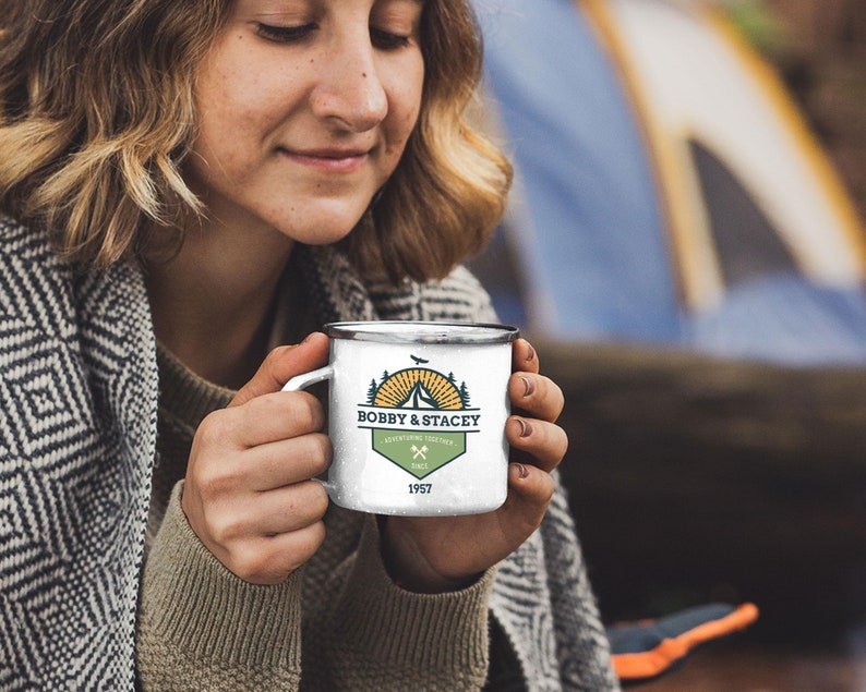 May include: White enamel camping mug with a silver rim. The mug features a green and gold design with the text "BOBBY & STACEY" and "ADVENTURING TOGETHER" with crossed axes and the year "1957". The mug is held by a person.