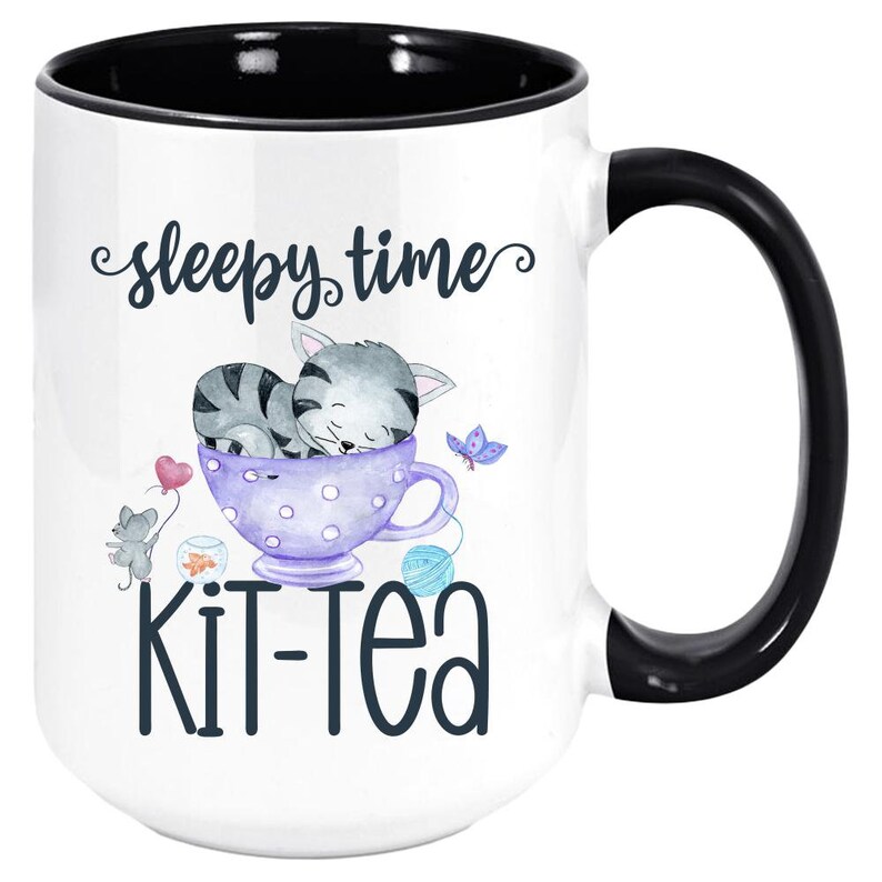 May include: White ceramic mug with a black interior and handle. The mug features a whimsical design with a sleeping gray cat in a purple teacup, the words "sleepy time" and "Kit-Tea". A mouse, butterfly, and yarn balls complete the design.