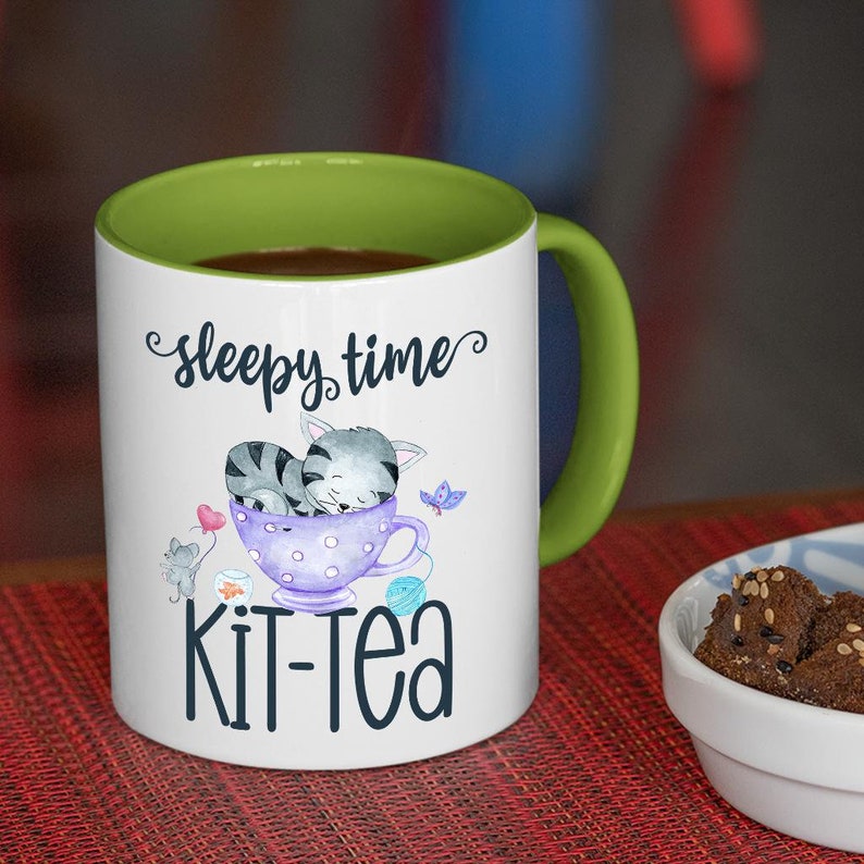 May include: White ceramic mug with a green interior and handle. The mug features the words "sleepy time" and "Kit-Tea" with a watercolor illustration of a sleeping gray cat in a teacup, a butterfly, and yarn.
