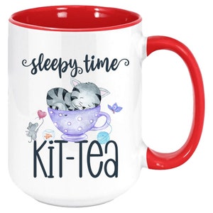 May include: White ceramic mug with a red interior and handle. The mug features a whimsical design with a sleeping gray tabby cat in a purple teacup, the words "sleepy time" and "kit-tea". A heart-shaped balloon and a butterfly are also included.
