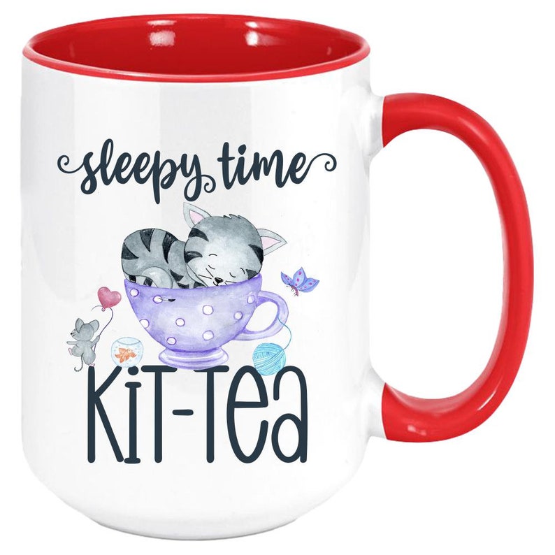 May include: White ceramic mug with a red interior and handle. The mug features a whimsical design with a sleeping gray tabby cat in a purple teacup, the words "sleepy time" and "kit-tea". A heart-shaped balloon and a butterfly are also included.