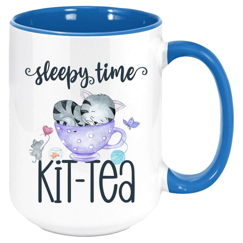 May include: White ceramic mug with a blue interior and handle. The mug features a whimsical design with a sleeping gray tabby cat in a teacup, the words "sleepy time" and "kit-tea". A mouse, butterfly, and yarn balls complete the design.
